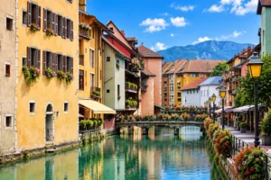 Vue d ensemble d un canal bordé de maisons pastel avec balcons fleuris, pont en pierre et montagnes à l horizon.