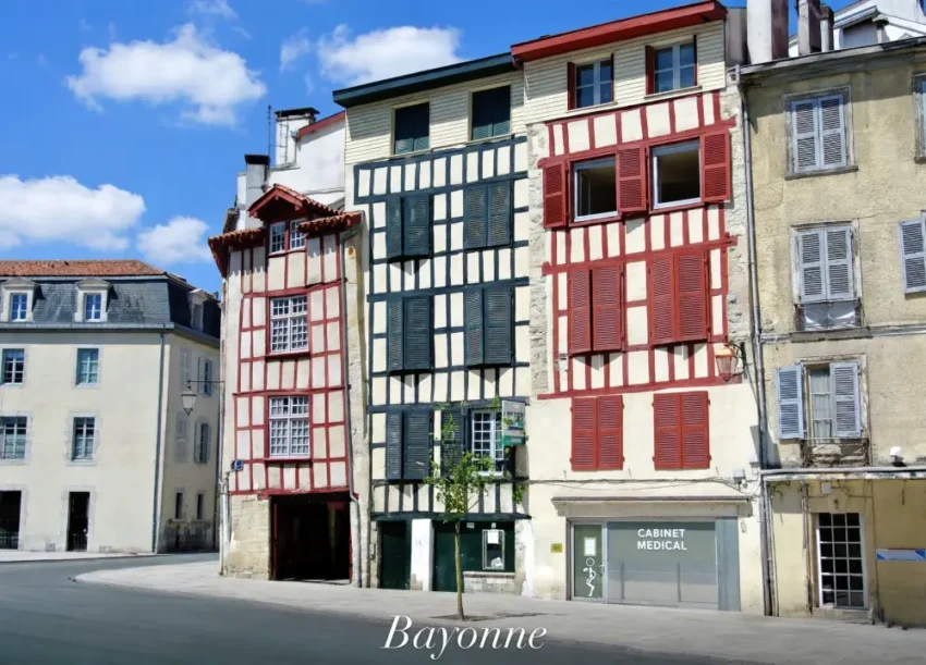 Immobilier à Bayonne en 2026 : le Pays basque est-il toujours inaccessible ? 4 Rangée de maisons à colombage colorées, cadres rouge et bleu, volets fermés ou ouverts, au rez-de-chaussée un cabinet médical, ciel bleu clair.