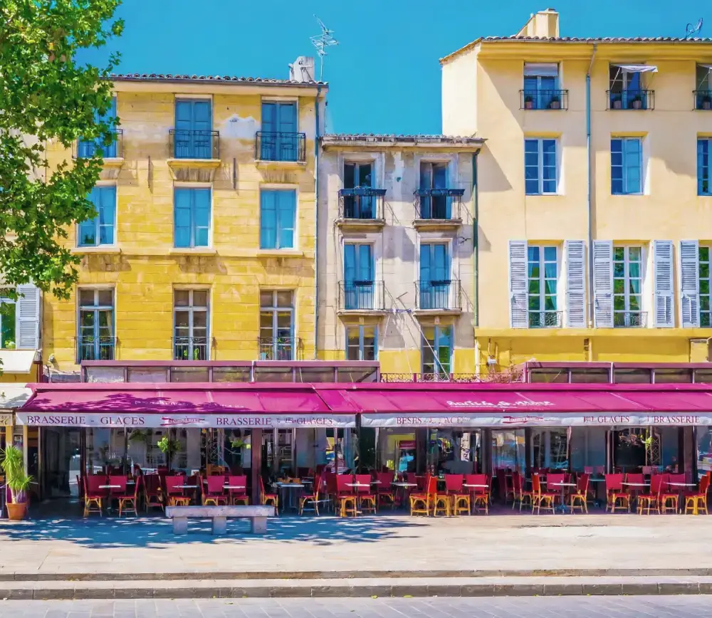 Trois facades jaune clair avec fenetres bleues et balcons en fer, devanture de brasserie avec chaises rouges sous un auvent rose, arbre a gauche.