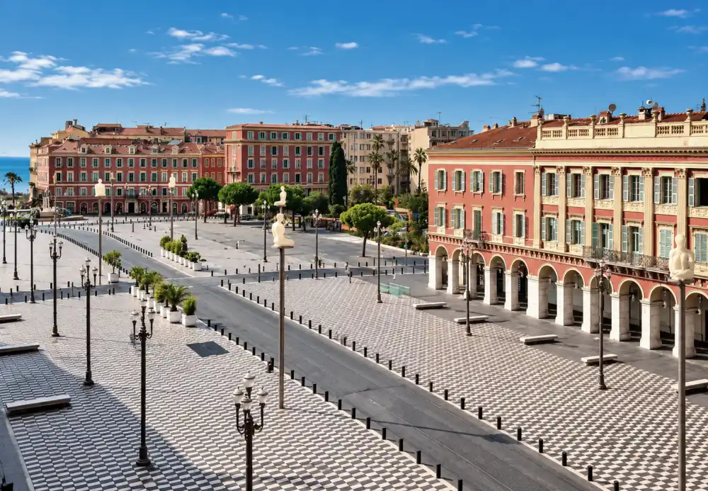 Immobilier à Nice en 2026 : jusqu’où les prix peuvent-ils encore monter ? 2 Vue en perspective d'une grande place pavée de carreaux noirs et blancs, des bâtiments roses à arches au bord, des lampadaires et statues.