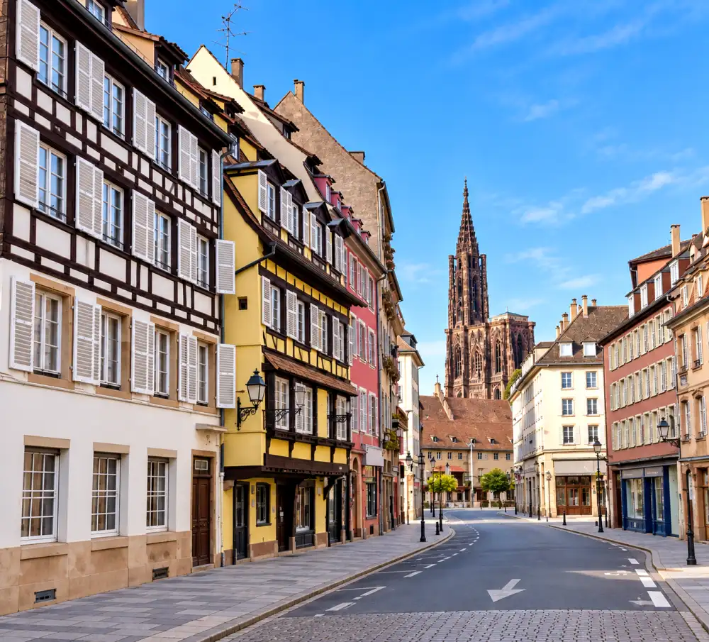 Vue d'une rue pavée bordée de maisons à colombage colorées, avec une grande cathédrale gothique au loin et un ciel bleu.