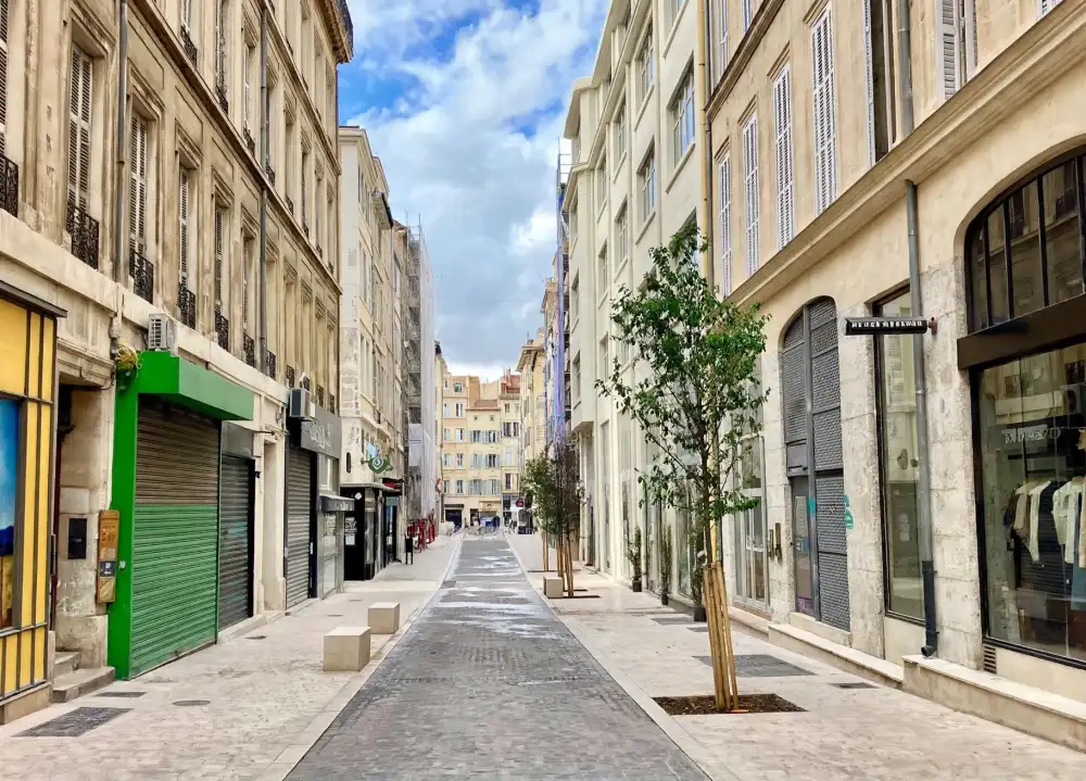 Immobilier à Marseille en 2026 : hausse des prix ou retournement du marché ? 7 vue d'une rue pietonne flanquee d'immeubles beige a façades anciennes, volets fermés, petits arbres et ciel partiellement nuageux.