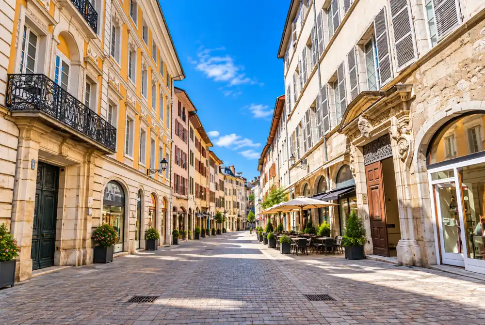Vue d'une rue piétonne bordée de façades claires, balcons en fer forgé, terrasses avec parasols et pavés sous un ciel bleu.