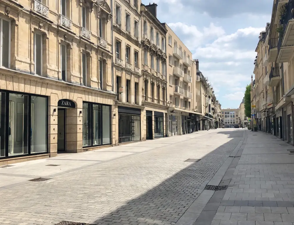vue en perspective rue pietonne bordees de facades en pierre beige avec magasin zara a gauche et peu de passants