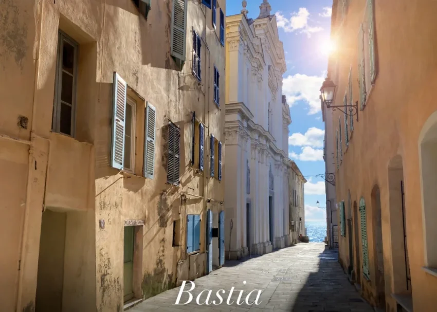 vue d'une ruelle pavée entre immeubles aux murs pastel et volets bleus, soleil à l'angle et mer au fond.