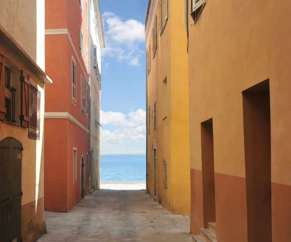Immobilier à Bastia en 2026 : le marché corse commence-t-il à ralentir ? 2 ruelle étroite entre deux immeubles aux murs orange et jaune, portes et volets, mer bleue au fond sous ciel clair