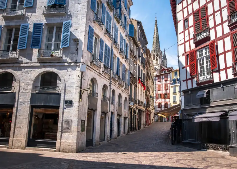 vue d une ruelle pavee entouree de batiments en pierre avec volets bleus et arches au rez-de-chaussee clocher au fond sous ciel bleu