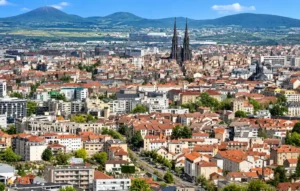 Vue aérienne d'une ville dense aux toits rouge orangé, une impressionnante cathédrale gothique au centre et des montagnes en arrière-plan.